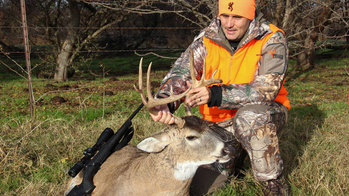 Hunter in orange vest holding buck by rack with black rifle propped up on deer