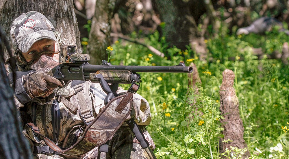 Turkey hunter leaning against tree pointing shotgun.