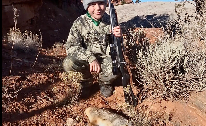 Young hunter in camo with rifle kneeling next to downed rabbit