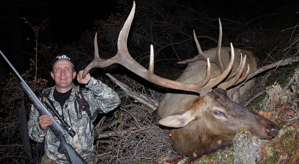 Male hunter holding rifle posing next to bull elk.