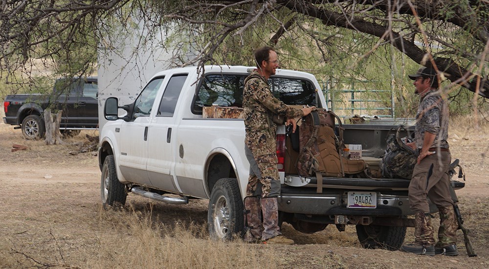 Two male hunters dressed in camouflage conversing while standing next to white Ford pickup truck.