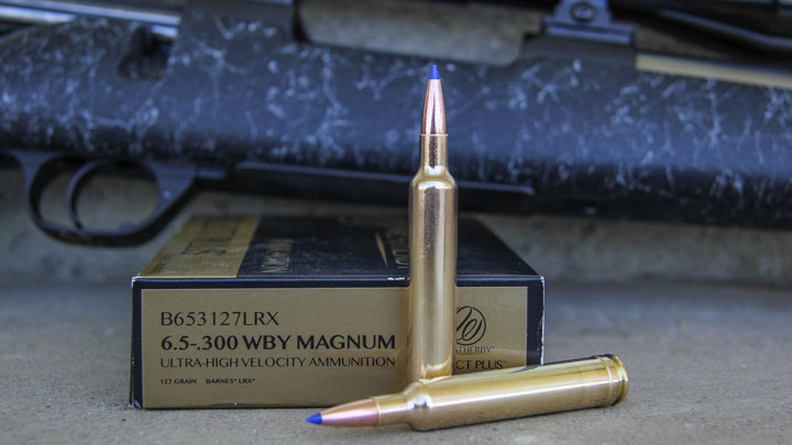 Two cartridges  sitting in front of a Weatherby box with a black bolt-action rifle in the background.