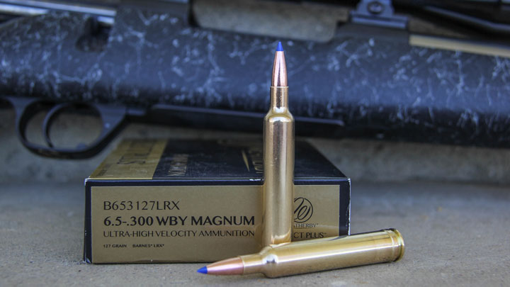 Two cartridges sitting in front of a Weatherby box with a black bolt-action rifle in the background.