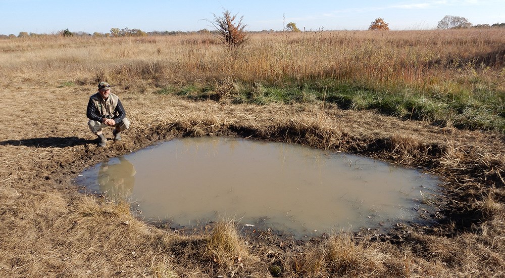 Man bending down next to small water hole in open field.