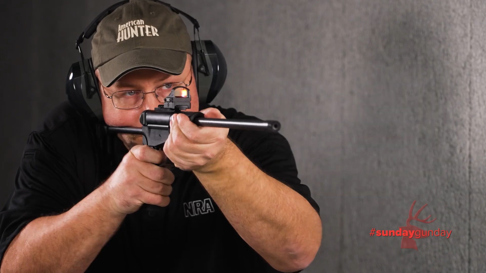Man wearing American Hunter ballcap earmuffs glasses facing camera shooting dark mountain arms stowaway rifle nra shirt indoors