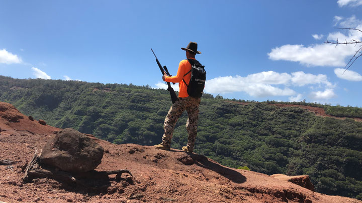 Hunter with a skyward rifle pressed to his thight stands, looking out over a cliff