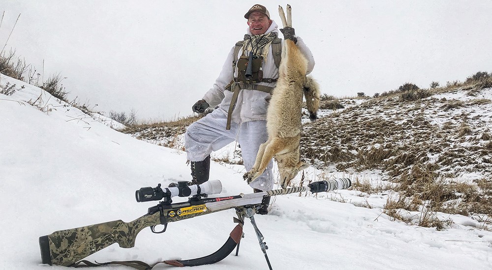 Male hunter holding up coyote on snowy mountainside.